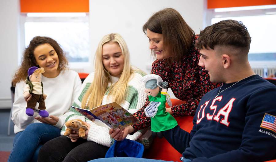 A person reads a children's book while three other people holding puppets and toys look on.
