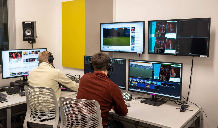 Two people sit at a desk. In front of them are several large screens showing a football match.