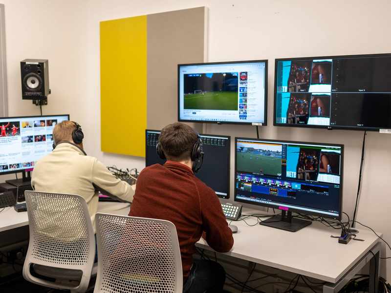 Two people sit at a desk. In front of them are several large screens showing a football match.