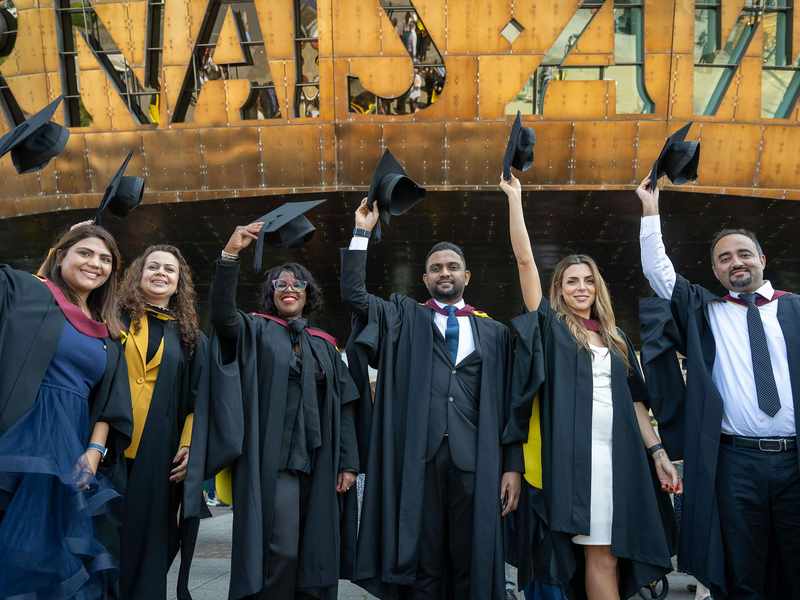 Six students in graduation gowns line up in front of the Wales Millennium Centre, holding their caps in the air.