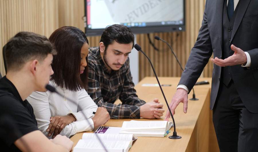 Three people sit at a desk in a courtroom. There are open textbooks in front of them.