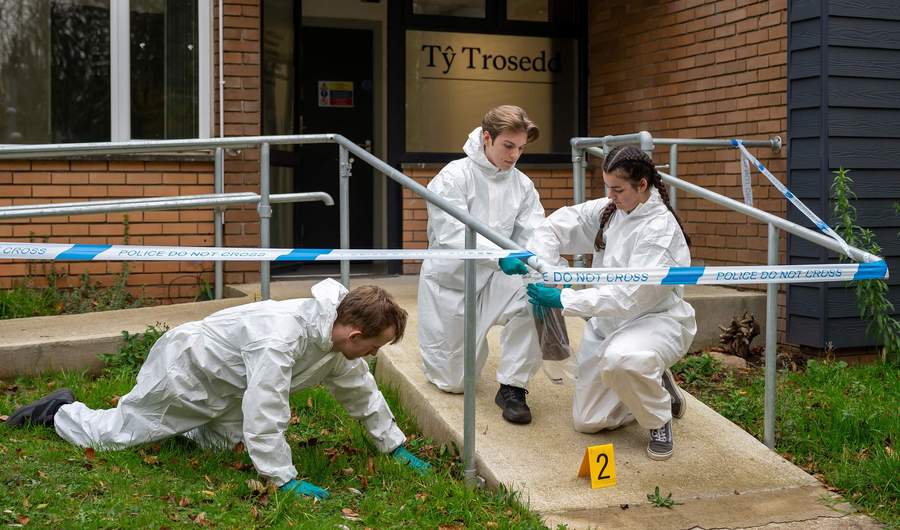 Three students wearing white jumpsuits and gloves inspect a fake crime scene for clues
