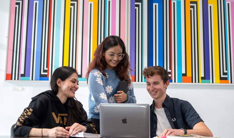 Two people sit at a table in front of a laptop. Behind them stands a third person.
