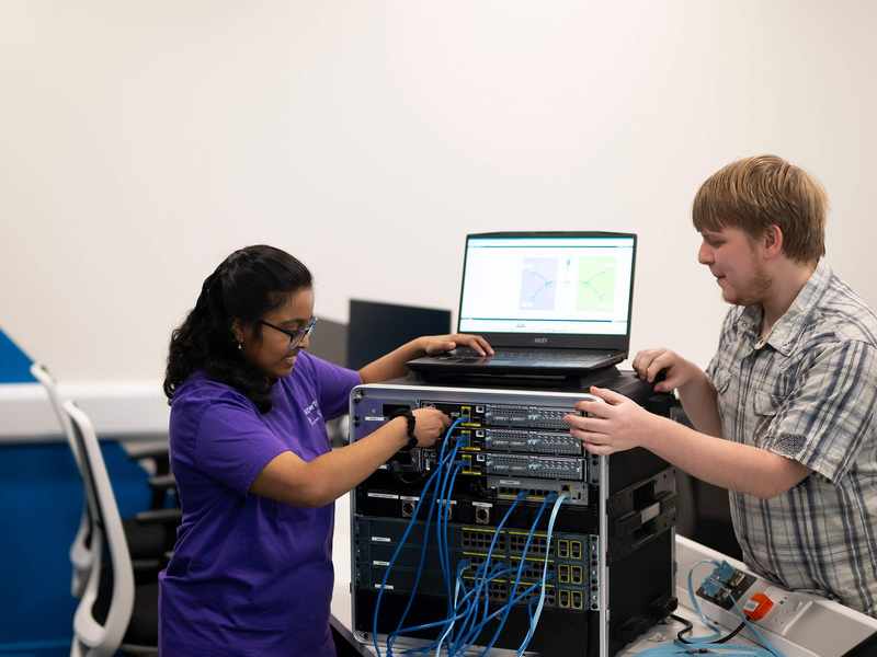 Two people plug cables into computer network equipment.