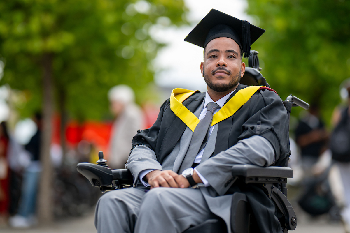 A man in a graduation gown and cap sits in a wheelchair, smiling proudly at his achievement.