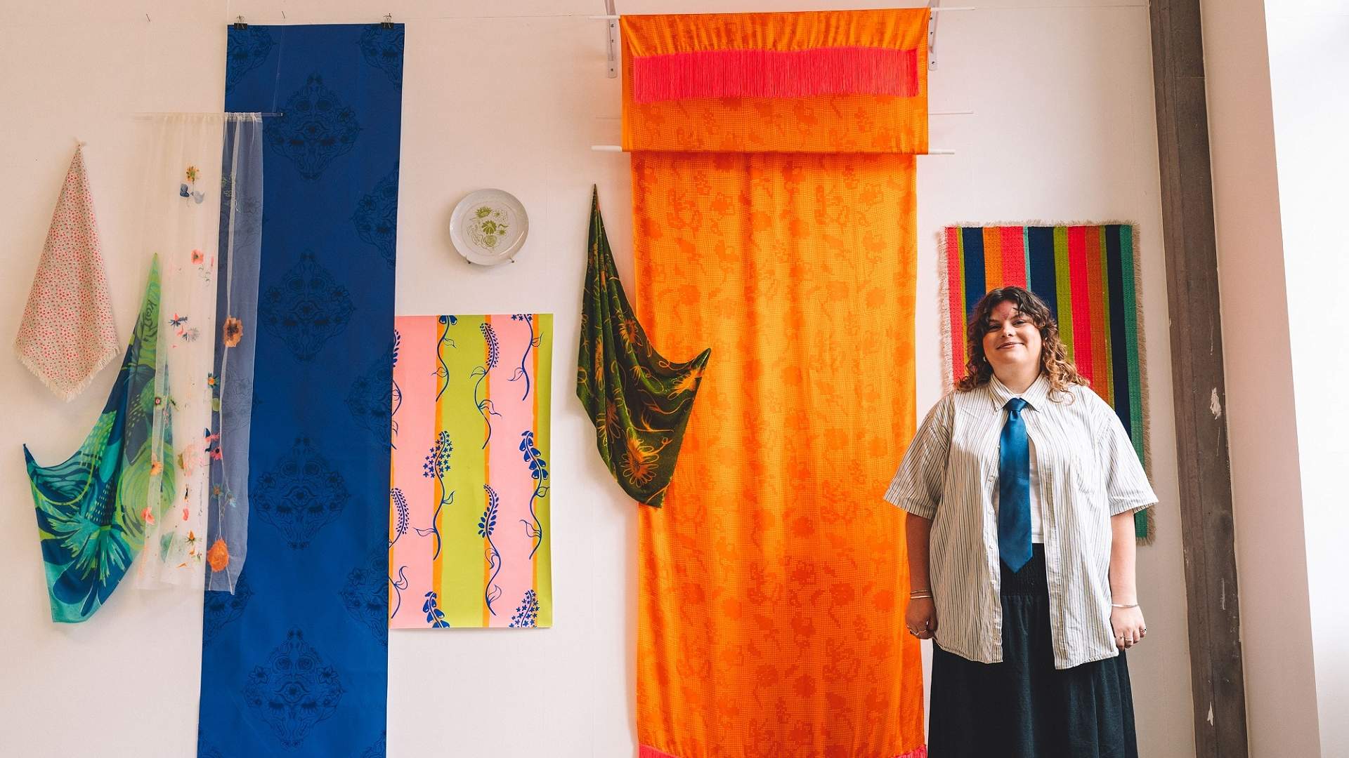 A young man in a shirt and tie stands beside a display of colourful textiles