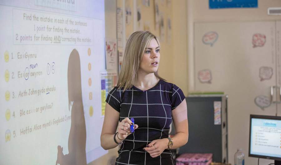 A teacher stands next to a whiteboard. Written on the whiteboard is some text in Welsh.