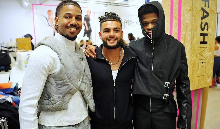 Three people pose arm in arm. In the background, costume sketches can been seen on the wall.