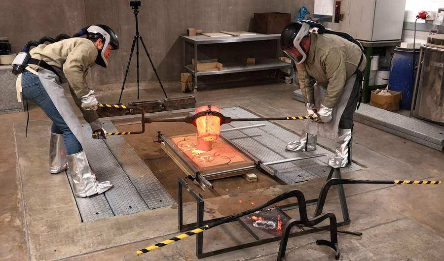 Two people wearing protective clothing pour molten metal into a mould