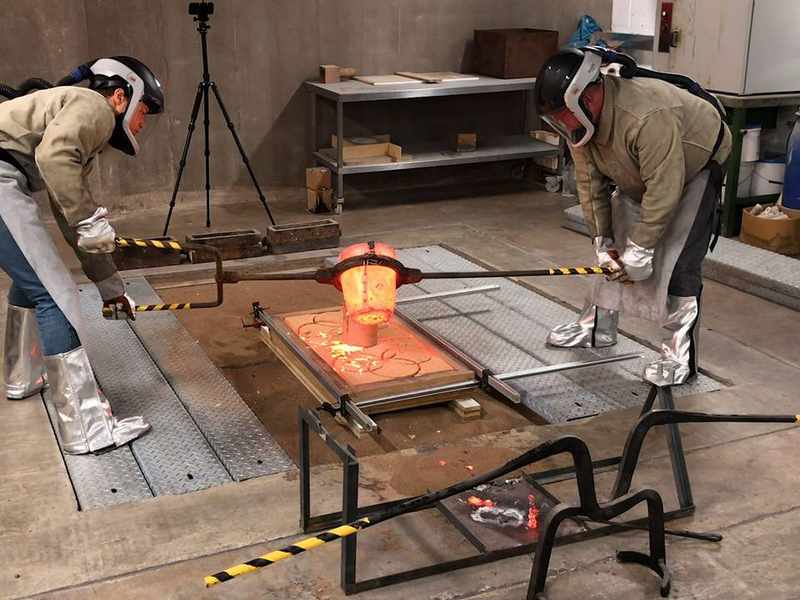 Two people wearing protective clothing pour molten metal into a mould
