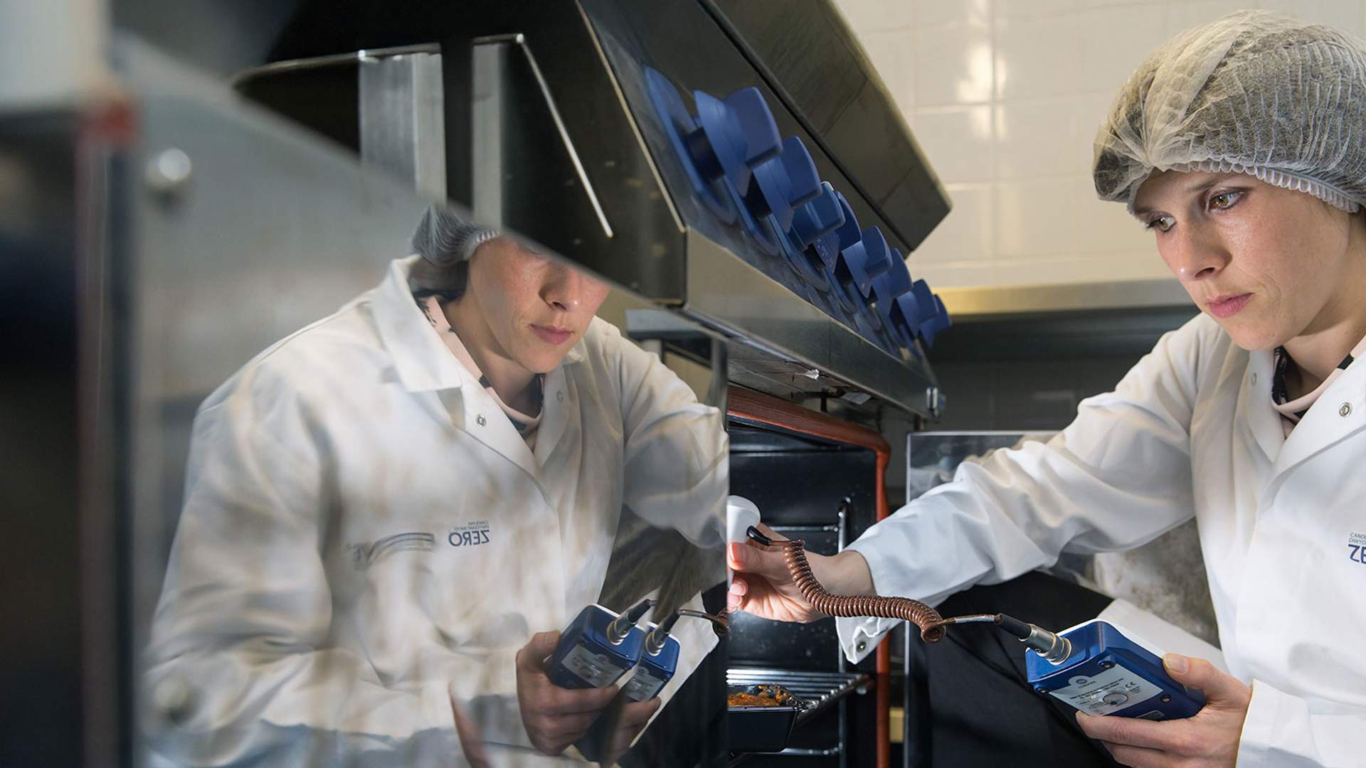 A person wearing a hairnet and a lab coat is inspecting equipment in a kitchen. They are holding a clipboard and appear focused on the task. A reflection of the scene is visible on a shiny surface.