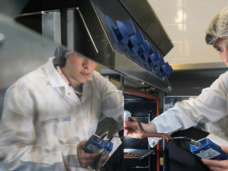 A person wearing a hairnet and a lab coat is inspecting equipment in a kitchen. They are holding a clipboard and appear focused on the task. A reflection of the scene is visible on a shiny surface.