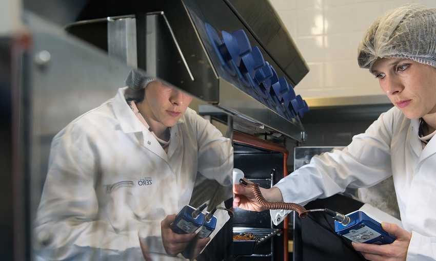 A person wearing a hairnet and a lab coat is inspecting equipment in a kitchen. They are holding a clipboard and appear focused on the task. A reflection of the scene is visible on a shiny surface.