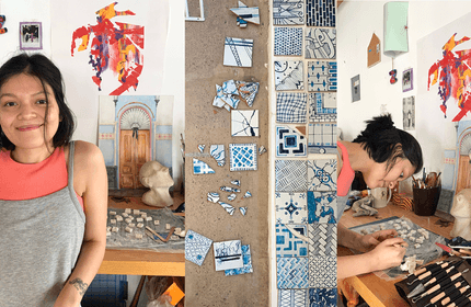 A woman stands and works at a table with mosaic pieces. Various small tiles are arranged on the table and a wall. Colorful artwork and a bust are visible in the background.
