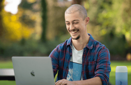 A person with a shaved head and a goatee is smiling while using a laptop outdoors. They are wearing a plaid shirt over a graphic tee. In the background, there is greenery and soft, natural light. A water bottle is visible on the table.