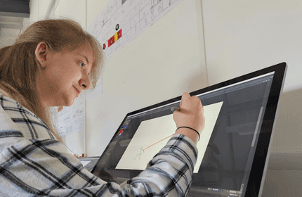 A person in a plaid shirt uses a stylus to draw on a large screen tablet at a desk. The screen displays a digital drawing interface. Wall charts with sketches are visible in the background.
