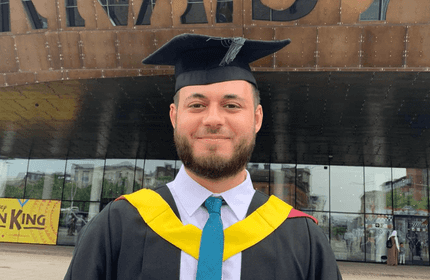 A person wearing a graduation cap and gown stands outside a building with large signage. The gown has yellow and red accents. A poster for The Lion King is partially visible in the background.