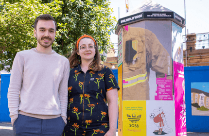 A man and woman stand smiling outdoors next to a kiosk covered in colorful posters. The woman wears a floral jumpsuit and headband, and the man wears a beige sweater. Trees and a building are visible in the background.