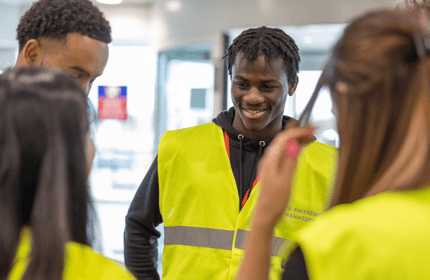 A group of people wearing bright yellow reflective vests are standing indoors, smiling and talking. One person in the center is looking at others, creating a friendly and engaging atmosphere.