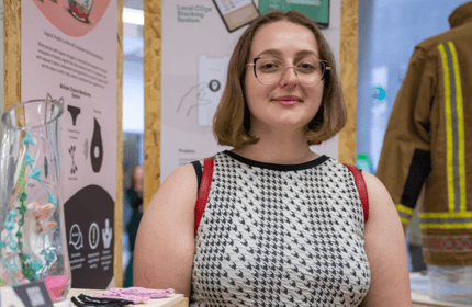 A person with shoulder-length hair and glasses stands smiling in front of a display booth. They are wearing a black-and-white patterned sleeveless top. Surrounding them are informational banners and a fire jacket, with other items visible on a table.