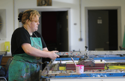 A person in a paint-splattered apron uses a screen printing press in a workshop. The scene includes paint containers and various tools. They appear focused on their task, with a partially open door and wall decorations in the background.