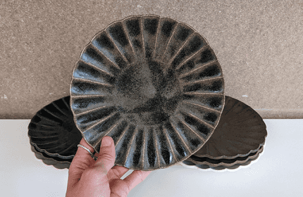 A hand holding a dark ceramic plate with a ribbed pattern. Several similar plates are stacked in the background on a white surface, against a neutral-colored wall.