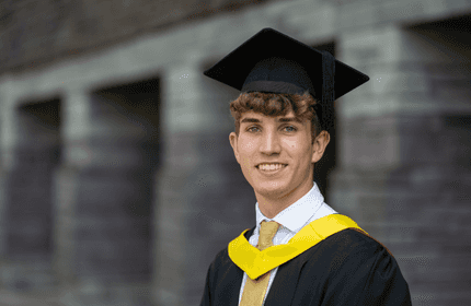 A young man in a graduation cap and gown stands smiling. He wears a white shirt, light brown tie, and has a yellow sash. The background is a stone wall in soft focus.